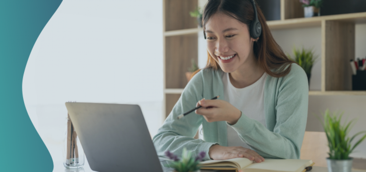 Image of an Asian woman wearing headphones, smiling and talking on a laptop at a desk.