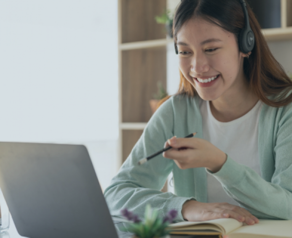 Image of an Asian woman wearing headphones, smiling and talking on a laptop at a desk.