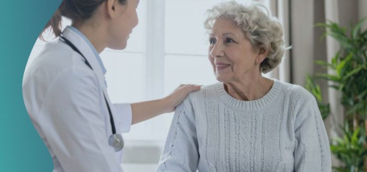 A health care professional in a white coat with a stethoscope places a hand on the shoulder of an older adult seated indoors during a medical consultation.