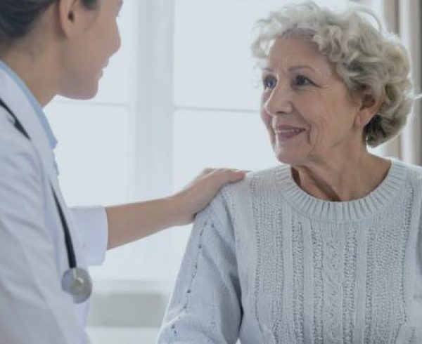 A health care professional in a white coat with a stethoscope places a hand on the shoulder of an older adult seated indoors during a medical consultation.