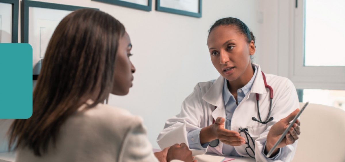 Healthcare provider in a white coat with a stethoscope explains information on a tablet to a seated patient during an office visit.