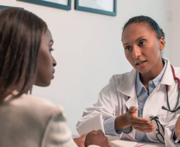 Healthcare provider in a white coat with a stethoscope explains information on a tablet to a seated patient during an office visit.
