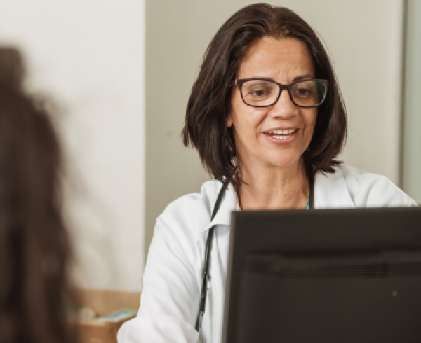 Healthcare professional wearing a stethoscope reviews information on a desktop computer while speaking with a patient seated across the desk in a clinical office setting.