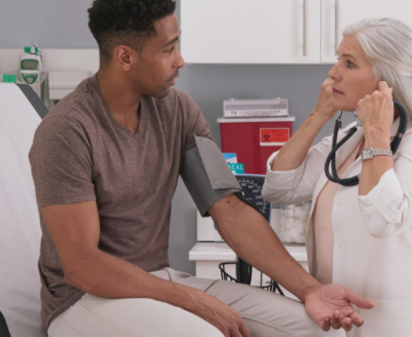 Healthcare professional using a stethoscope and blood pressure cuff to check a patient's blood pressure during a primary care appointment in a medical exam room. The patient is seated on an examination table while receiving a routine preventive health screening.